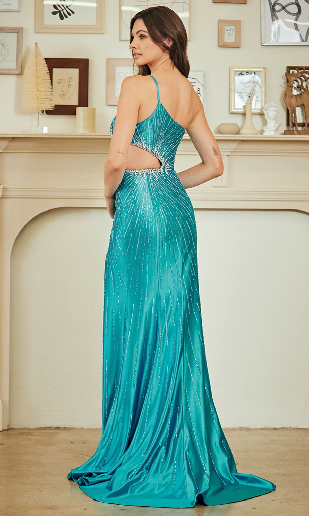 A woman wearing the Amelia Couture SU097, a teal, floor-length prom dress with a high leg slit and cutout back, stands by a fireplace adorned with small framed art and ornaments.